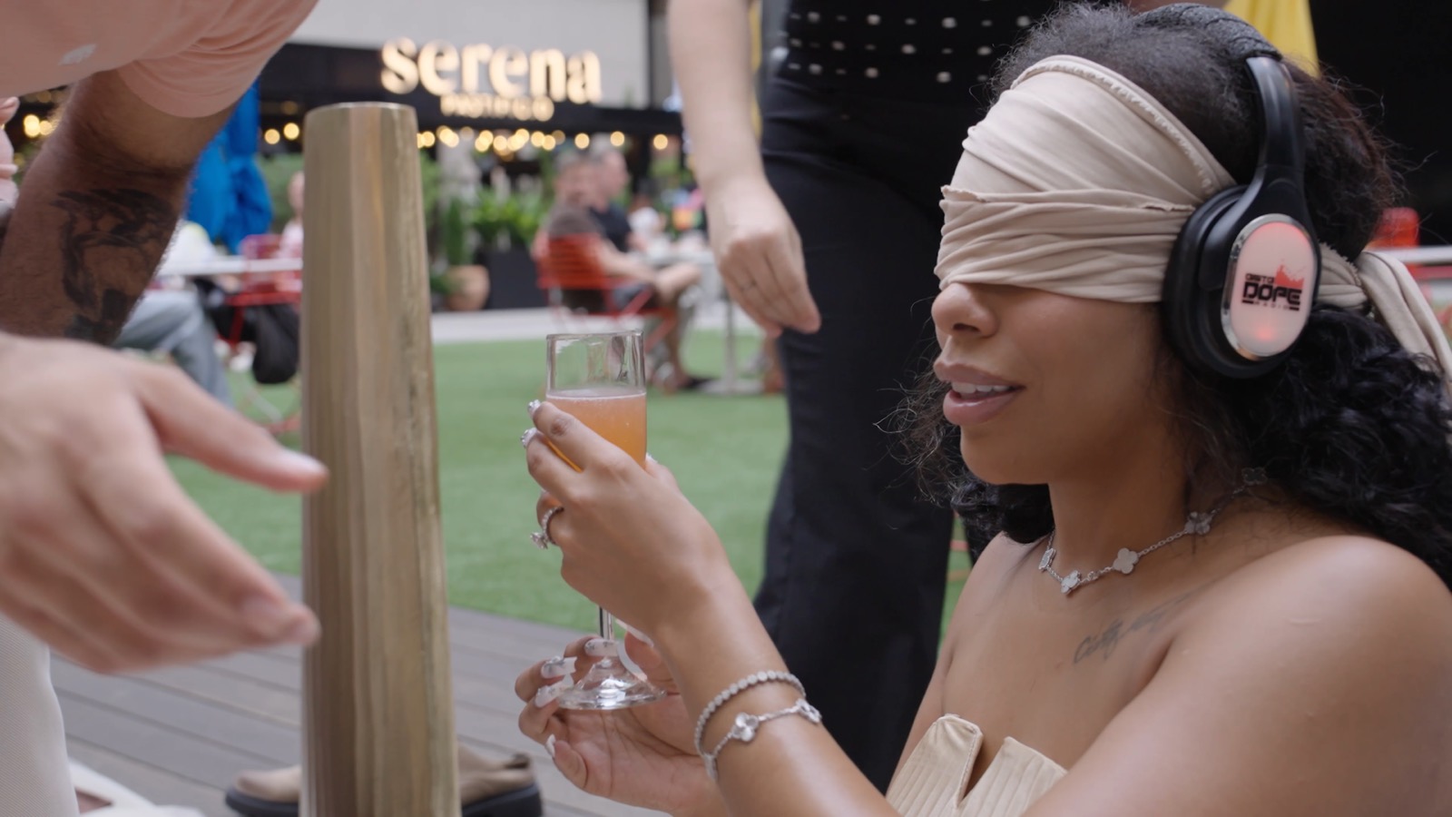 A guest wearing a blindfold and headphones raises a coupe glass at the table.